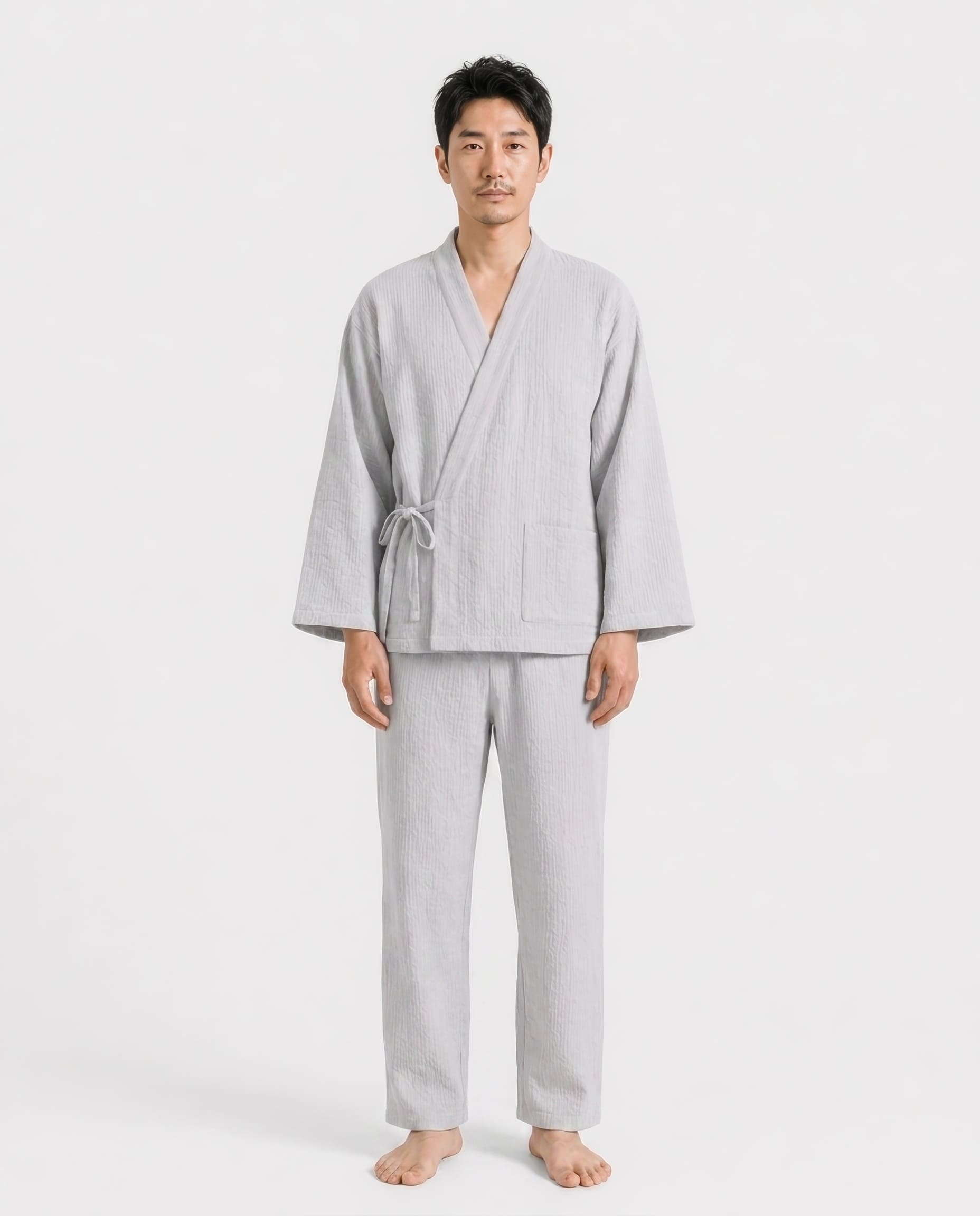 A man wearing a light gray two-piece pajama set consisting of a long-sleeve wrap robe with a drawstring and matching relaxed pants, standing against a plain light gray studio background.