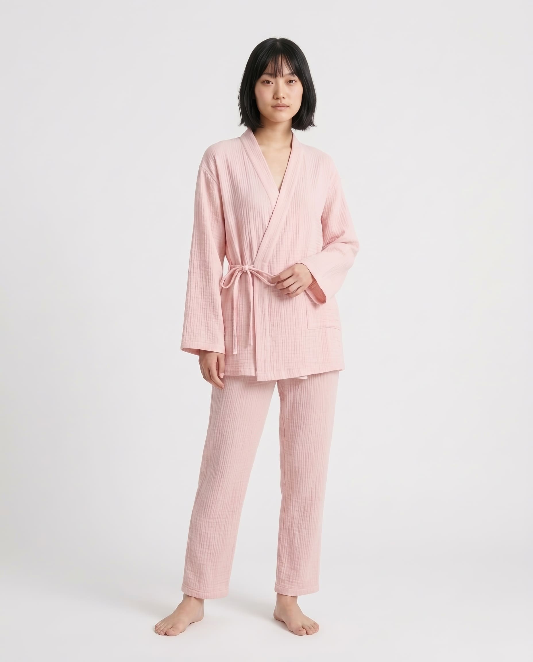 A woman modeling a light pink two-piece pajama set with a textured waffle fabric, consisting of a long-sleeve tie-front robe and matching pants, against a plain light gray studio background.